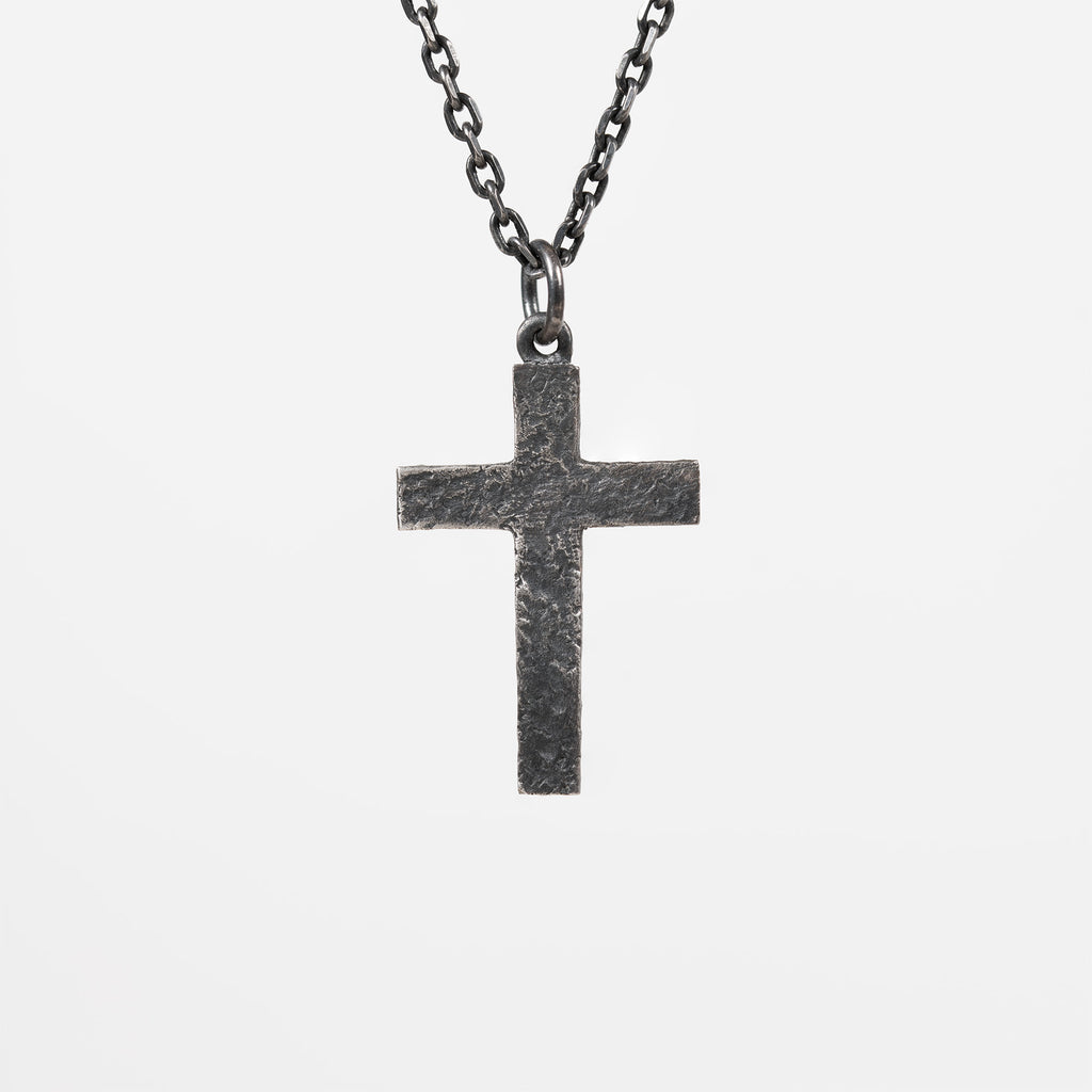 Back of the oxidized silver cross, plain with a rough carved texture and no figure or stone, hanging from a dark chain against light gray