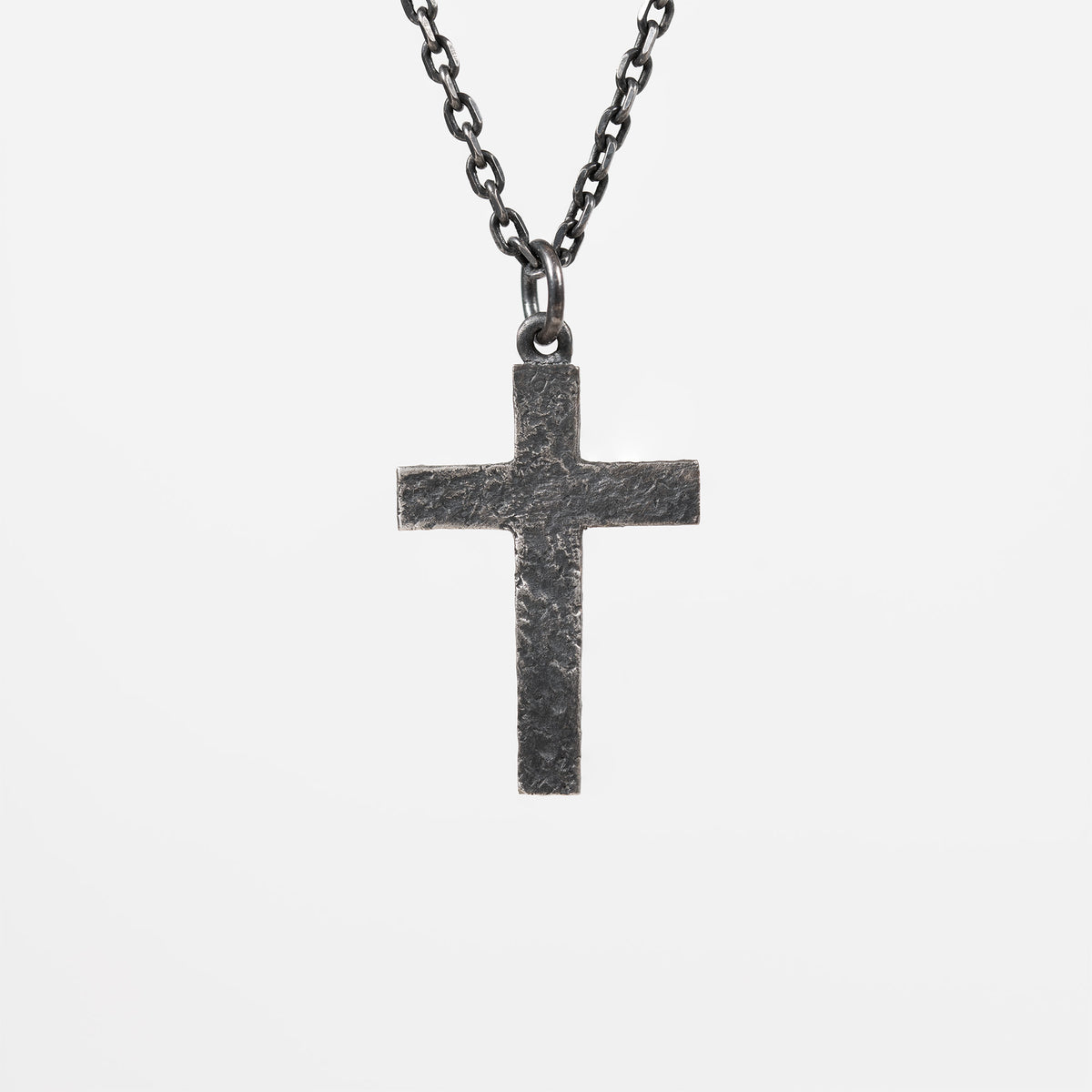 Back of the oxidized silver cross, plain with a rough carved texture and no figure or stone, hanging from a dark chain against light gray