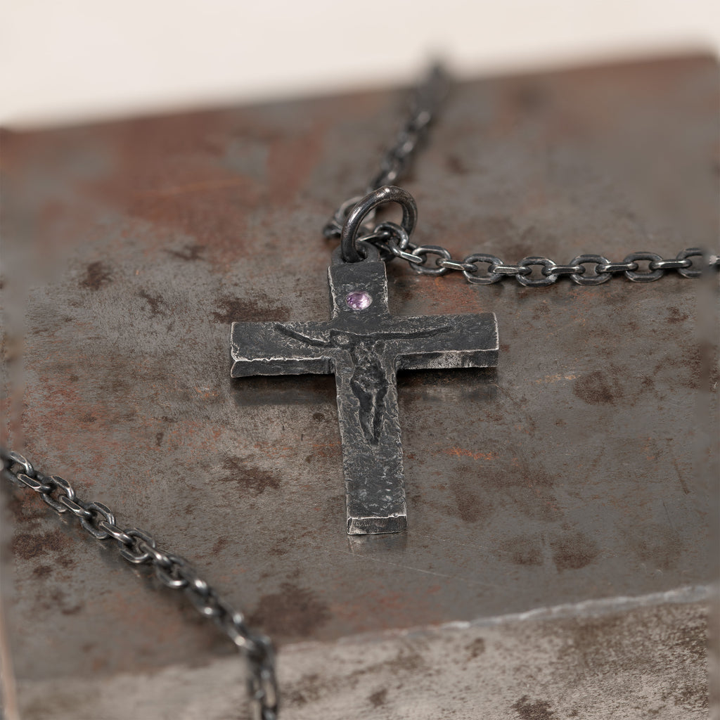 Carved crucifix with small purple cubic zirconia lying on a rusted metal block, its dark chain looping across the block