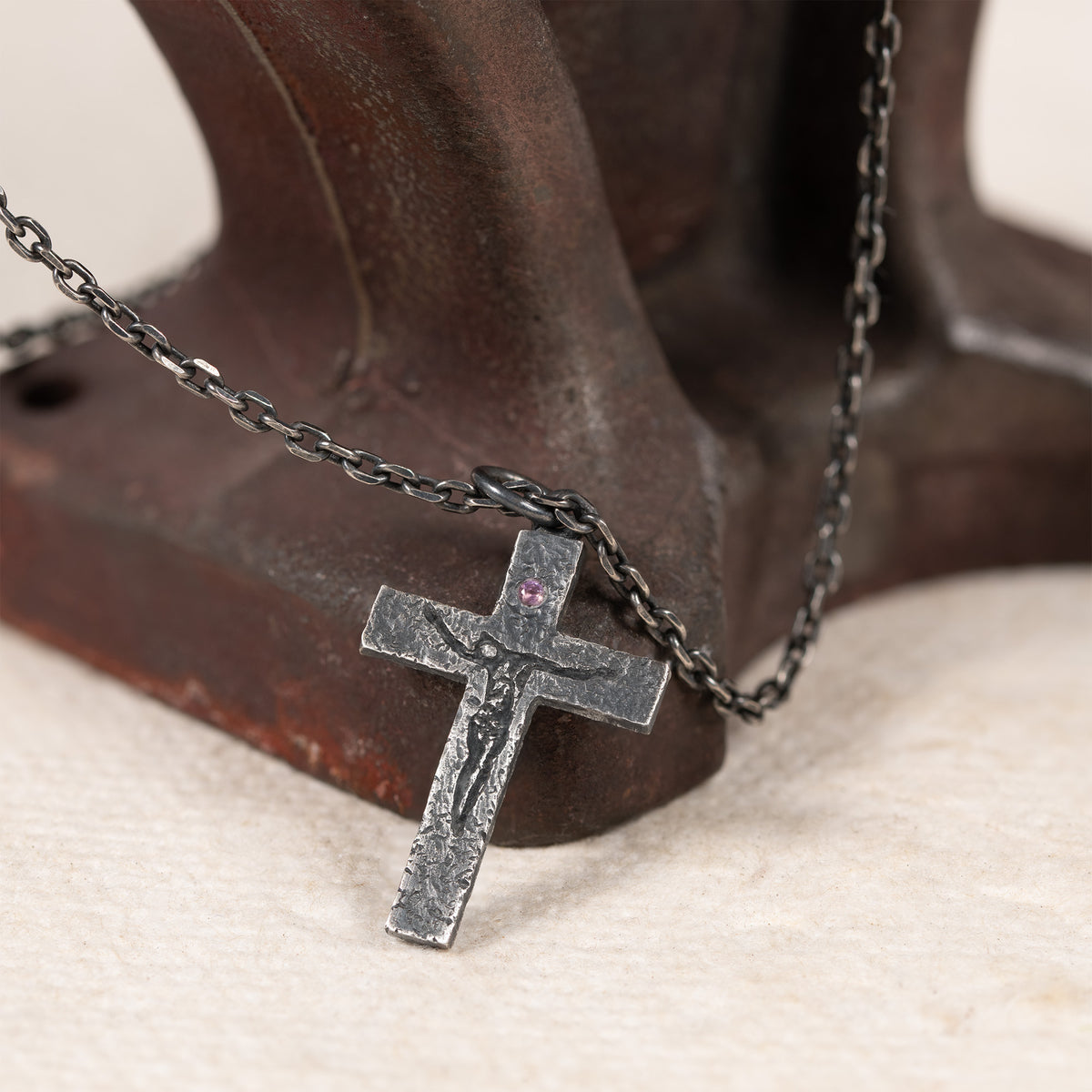 Crucifix with pink-purple cubic zirconia leaning against an old dark red metal stand, its chain draped over the stand on cream fabric