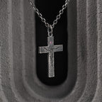 Oxidized crucifix pendant on a dark chain, framed by a gray concrete arch with a black opening behind it