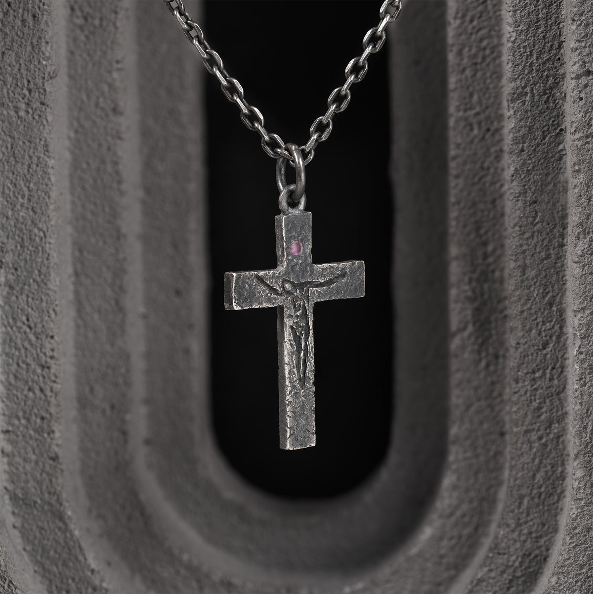 Oxidized crucifix pendant on a dark chain, framed by a gray concrete arch with a black opening behind it