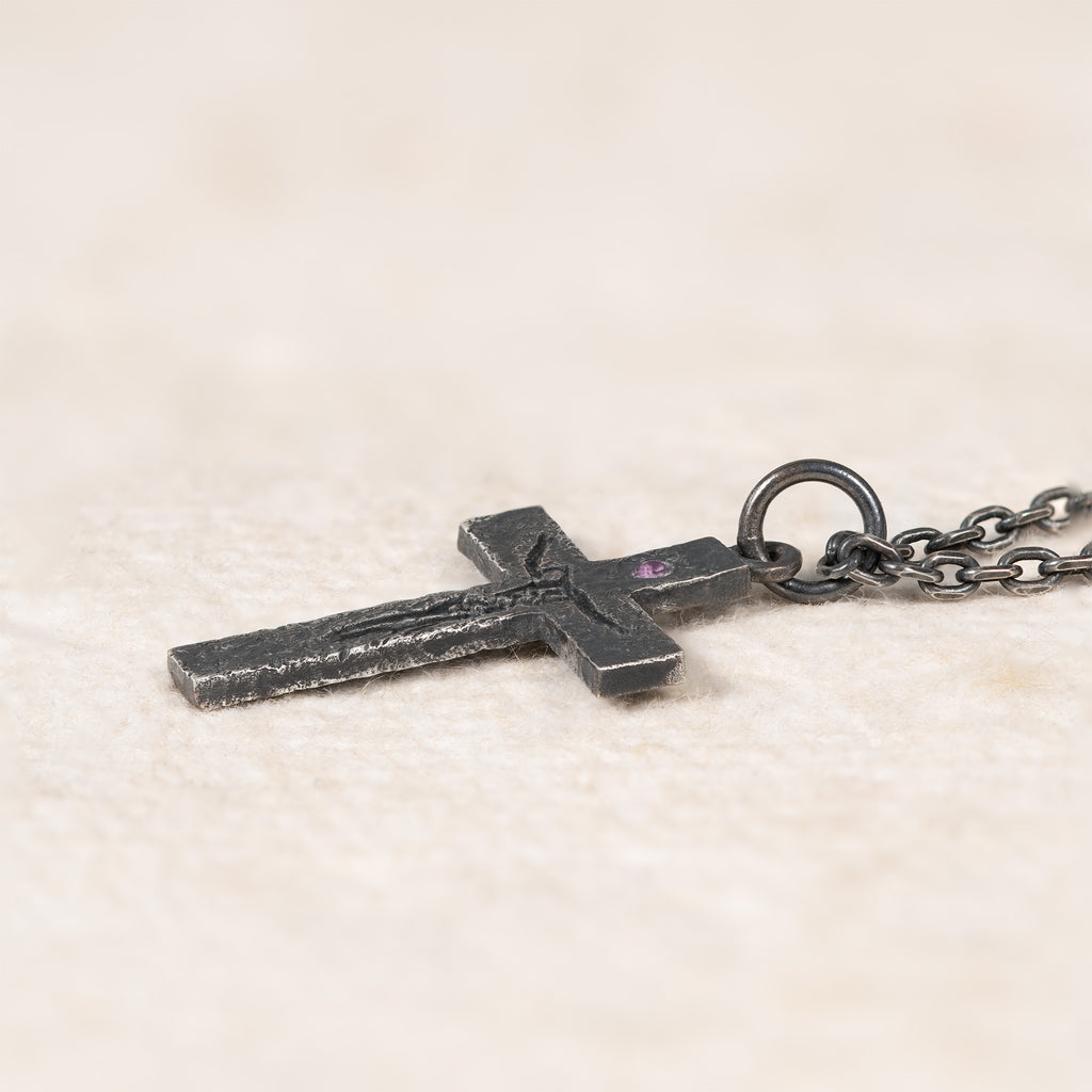 Side view of the cross lying on cream fabric, showing its thin profile, the raised carving and the purple cubic zirconia setting next to the bail