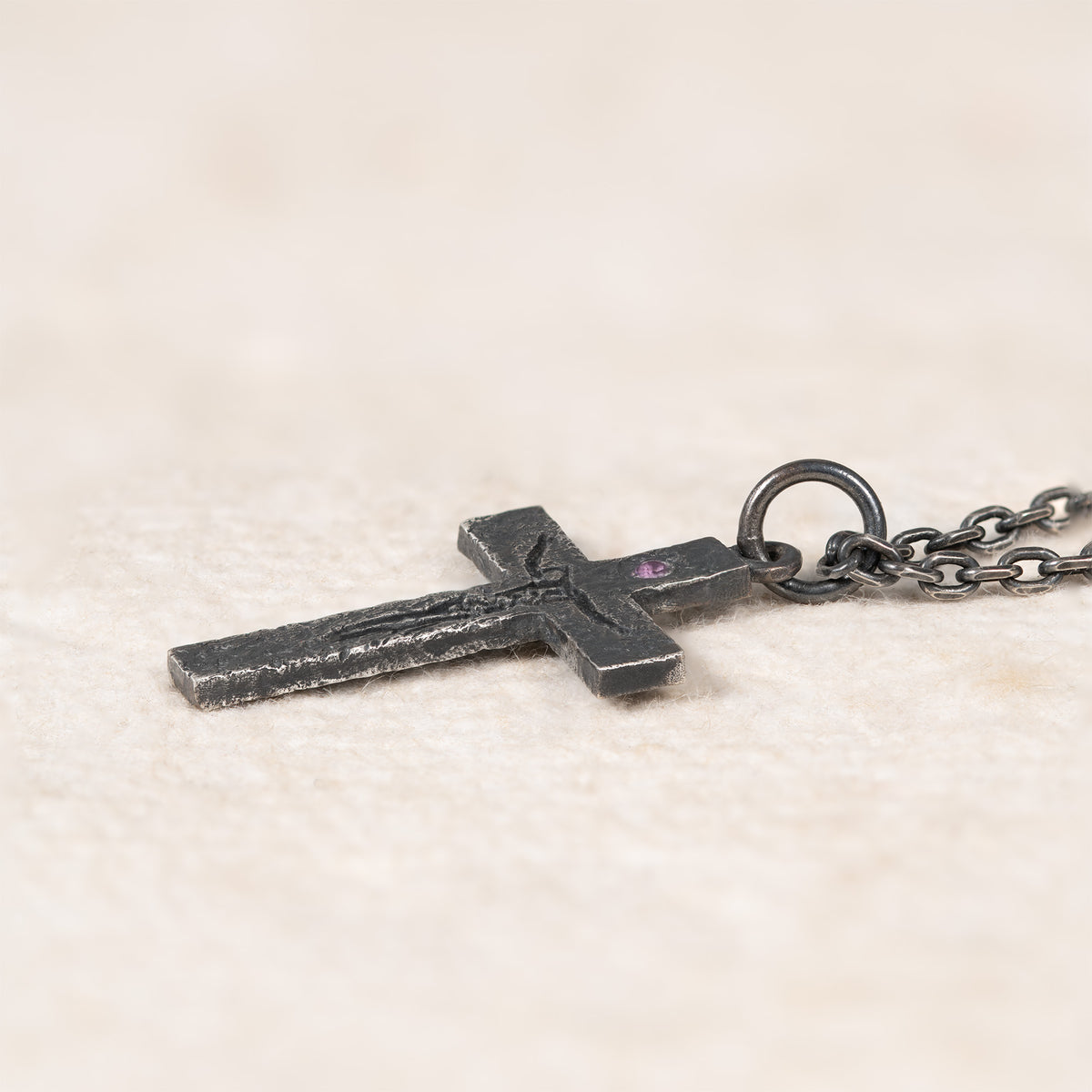 Side view of the cross lying on cream fabric, showing its thin profile, the raised carving and the purple cubic zirconia setting next to the bail