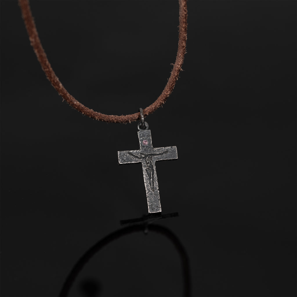 Crucifix pendant on a brown cord against a black reflective surface, with its reflection showing below