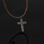 Crucifix pendant on a brown cord against a black reflective surface, with its reflection showing below