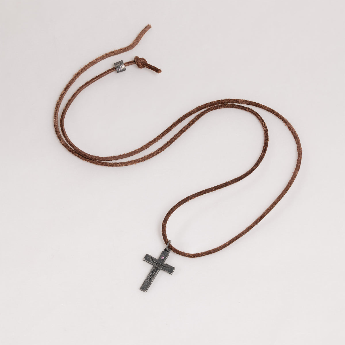 Crucifix pendant on a long brown cord with a small metal bead and knot near the cord ends, laid flat on a white background