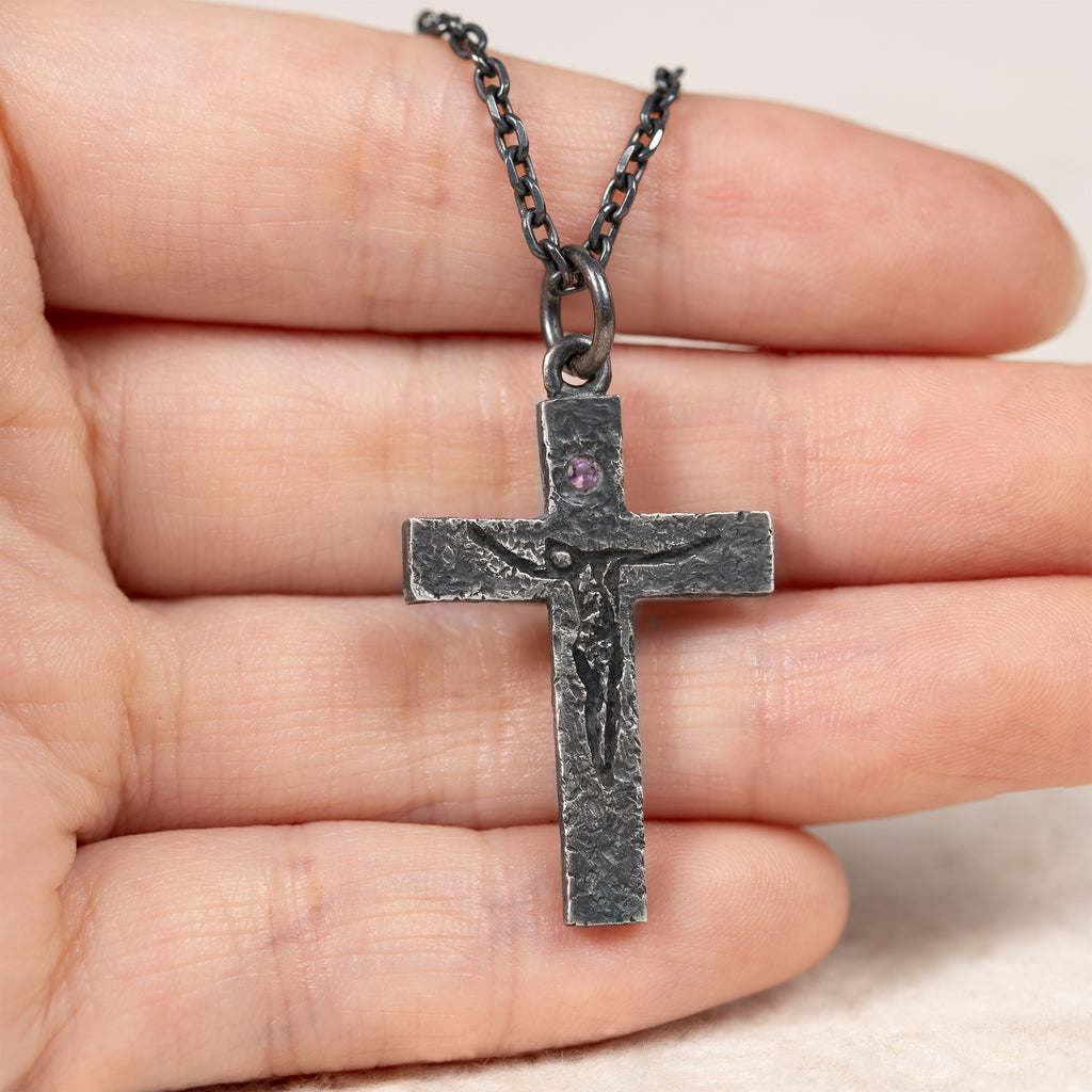 Crucifix pendant resting across the fingers of an open hand to show its size, with the purple cubic zirconia and carved figure facing up