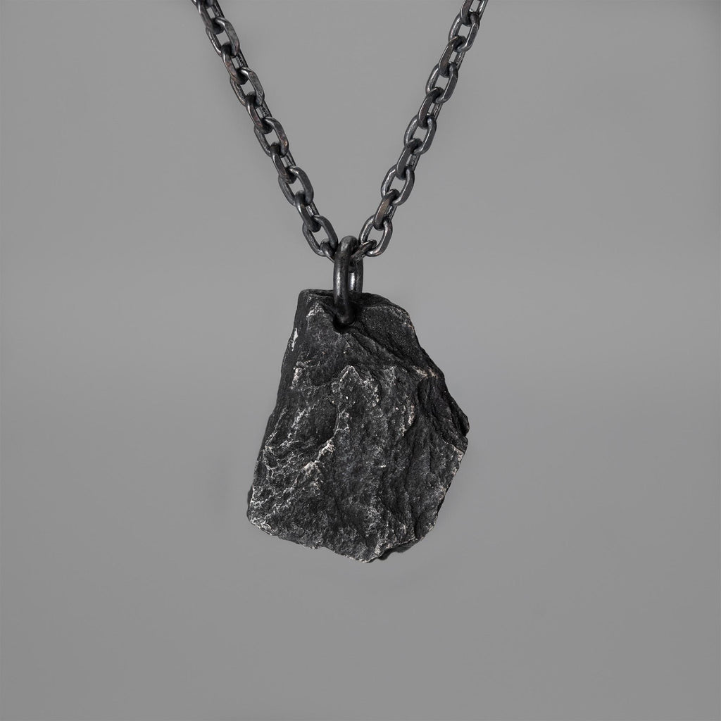 A dark, rugged silver pendant shaped like a natural rock, hanging from a black oxidized chain against a plain grey background. The pendant has a raw, textured surface with visible ridges and natural contours.