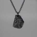 A dark, rugged silver pendant shaped like a natural rock, hanging from a black oxidized chain against a plain grey background. The pendant has a raw, textured surface with visible ridges and natural contours.