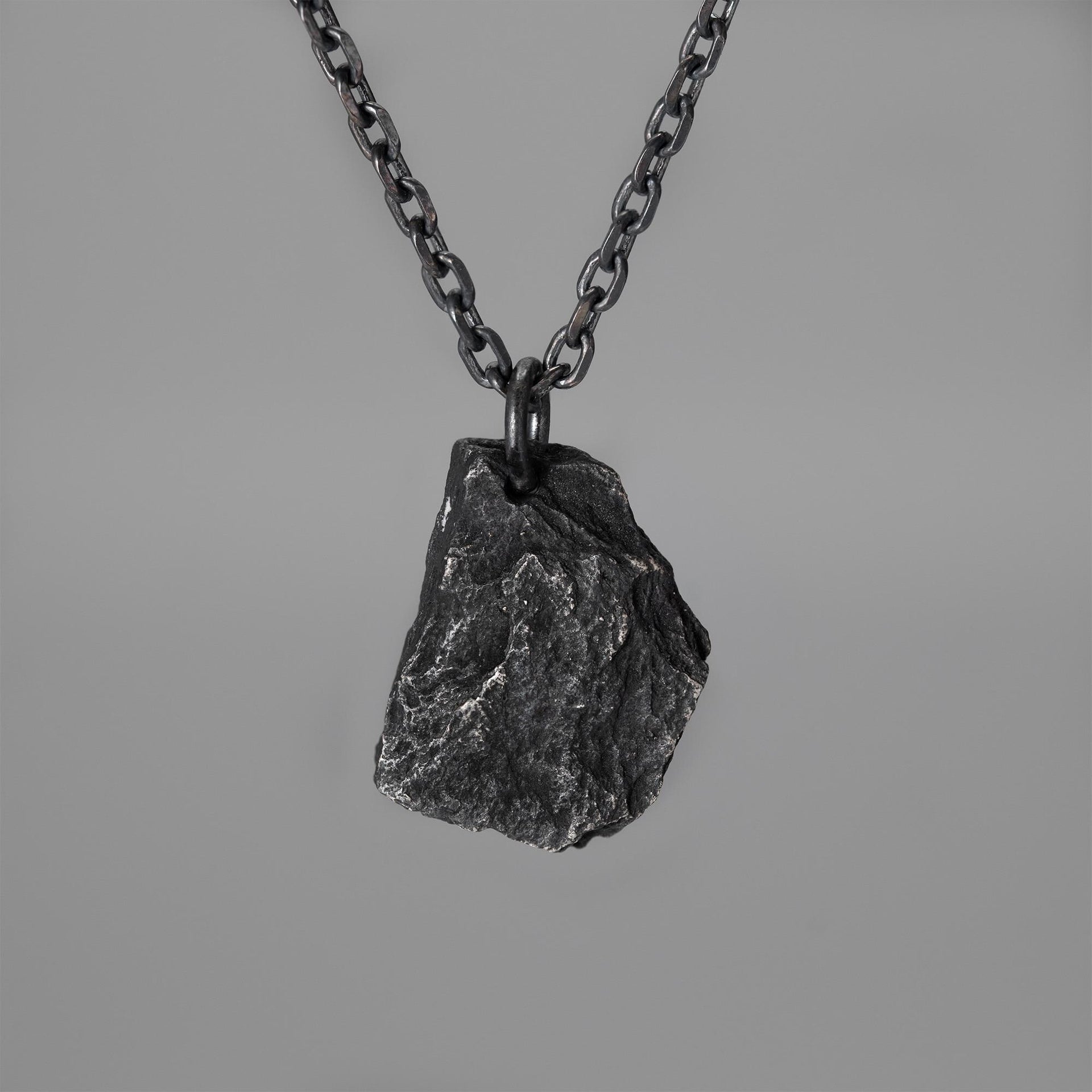 A dark, rugged silver pendant shaped like a natural rock, hanging from a black oxidized chain against a plain grey background. The pendant has a raw, textured surface with visible ridges and natural contours.