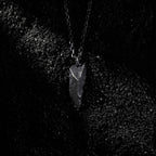 A dark, rugged silver pendant with a pointed, angular shape and rough texture, resting on a bed of black gravel. The pendant hangs from a black oxidized chain, highlighted by dramatic side lighting against the textured surface.
