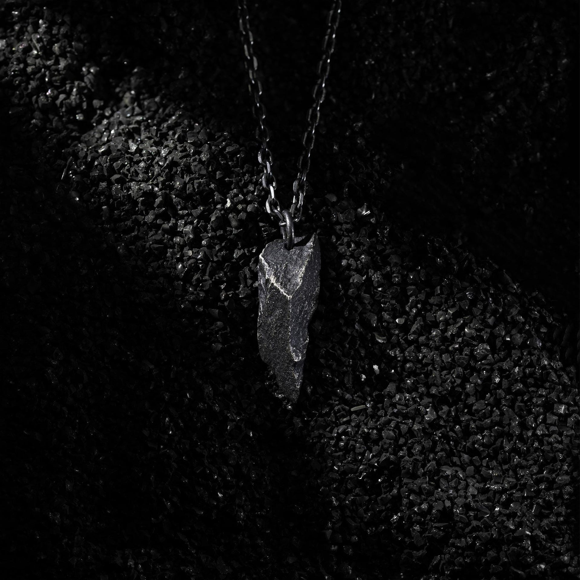 A dark, rugged silver pendant with a pointed, angular shape and rough texture, resting on a bed of black gravel. The pendant hangs from a black oxidized chain, highlighted by dramatic side lighting against the textured surface.