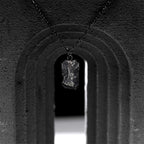 A dark, rugged silver pendant with a rough, chiseled surface hangs from a black oxidized chain, suspended in the center of a textured, arched black stone structure.