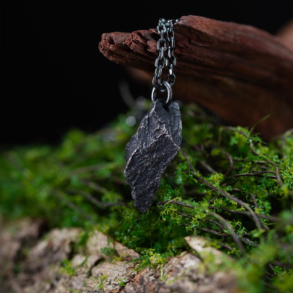 A dark, rugged silver pendant with a pointed, rock-like shape and textured surface, hanging from a black oxidized chain. The pendant rests against driftwood and green moss, evoking a natural forest setting.