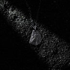 A dark, rugged silver pendant shaped like a natural rock, lying on a bed of black gravel under a dramatic beam of light. The pendant hangs from a black oxidized chain, with its textured surface catching subtle highlights.