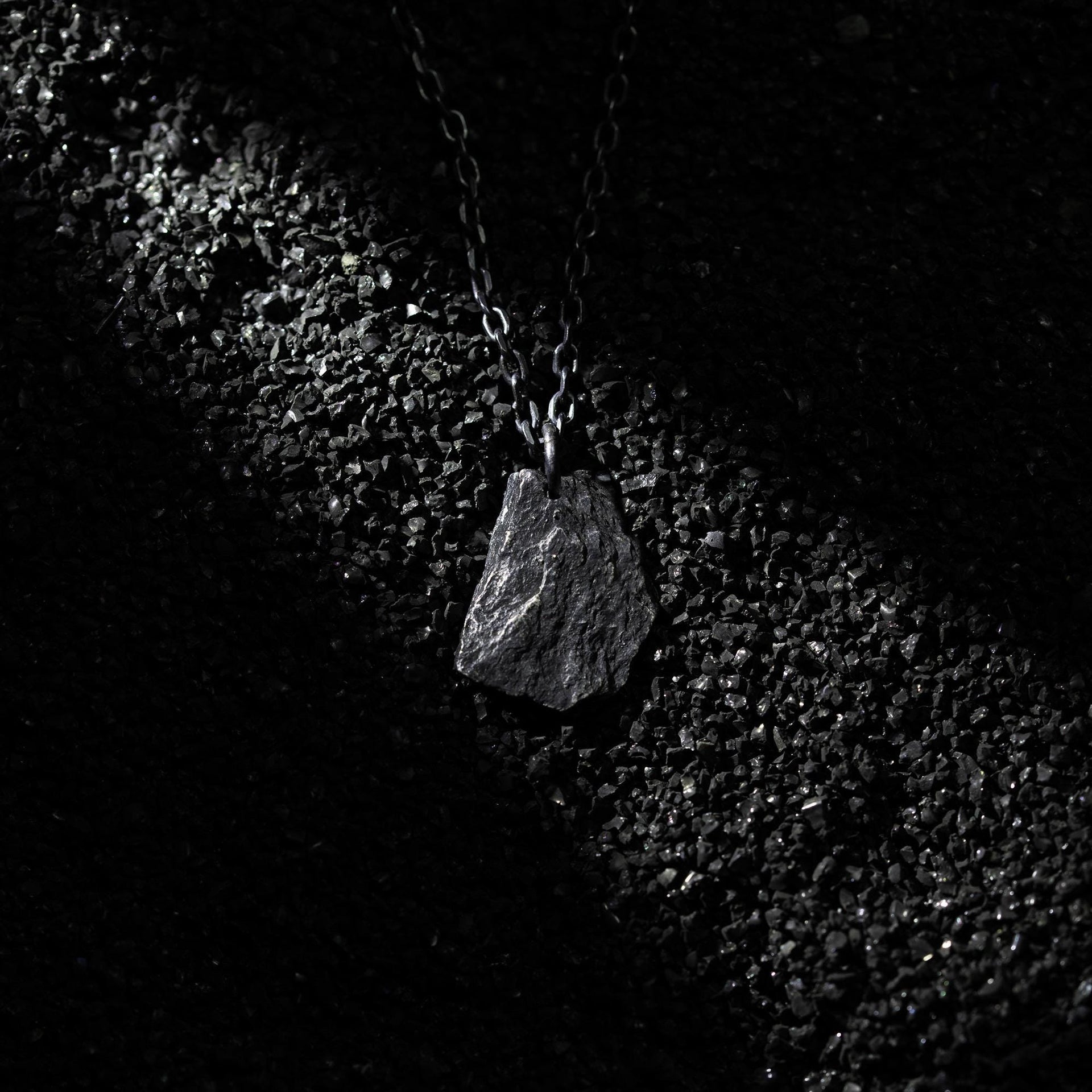 A dark, rugged silver pendant shaped like a natural rock, lying on a bed of black gravel under a dramatic beam of light. The pendant hangs from a black oxidized chain, with its textured surface catching subtle highlights.