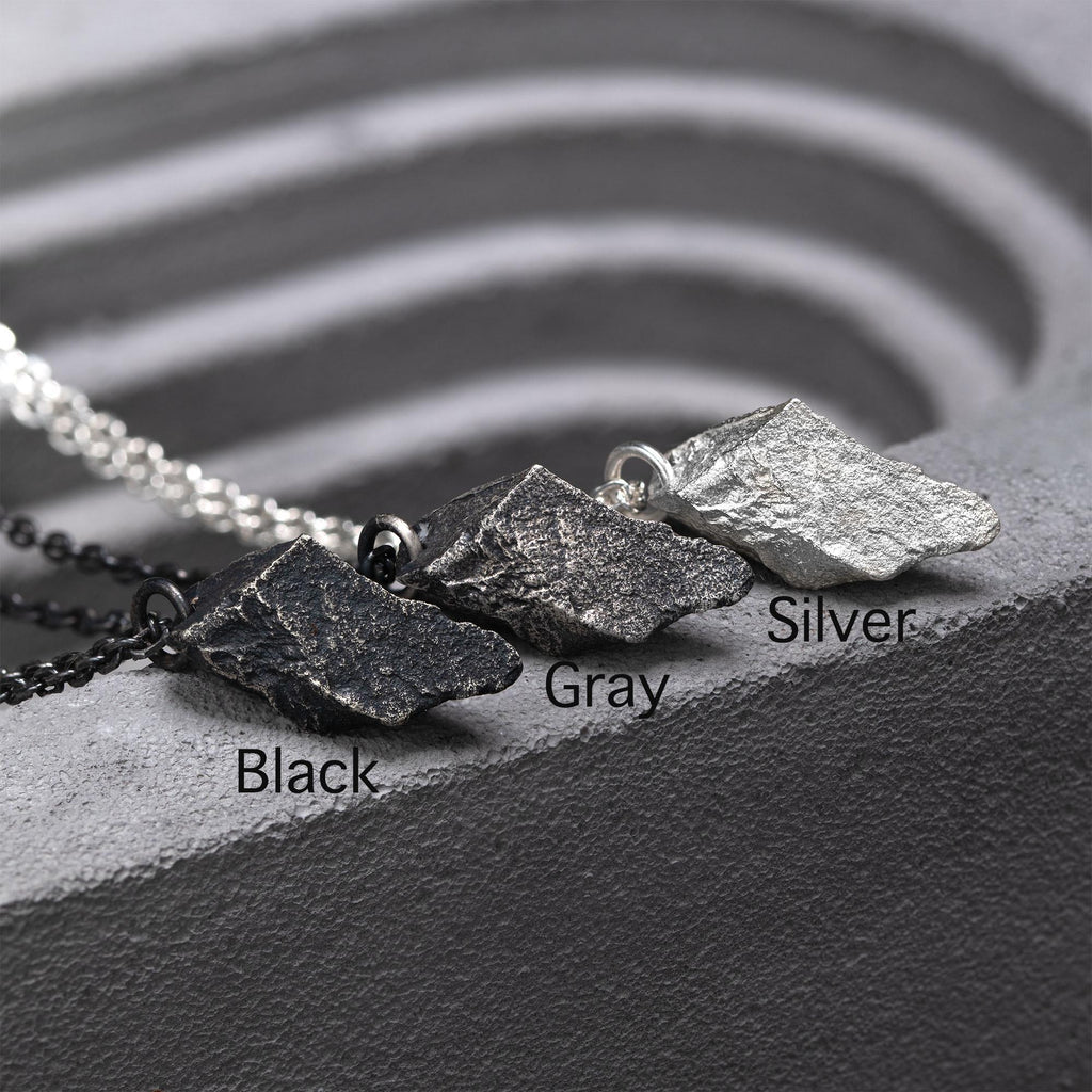 Three rugged silver pendants with angular, rock-like textures displayed side by side on a gray sculpted surface. The finishes are labeled Black, Gray, and Silver, each paired with matching chains.