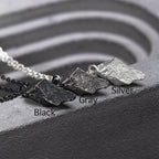 Three rugged silver pendants with angular, rock-like textures displayed side by side on a gray sculpted surface. The finishes are labeled Black, Gray, and Silver, each paired with matching chains.
