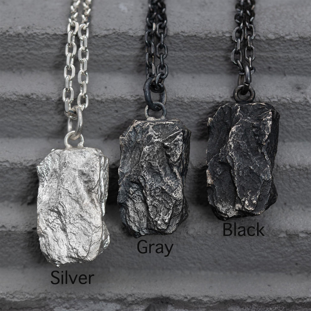 Three rugged silver pendants with raw, layered textures hanging side by side on chains, displayed against a gray textured background. The finishes shown are labeled Silver, Gray, and Black from left to right.