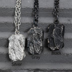 Three rugged silver pendants with raw, layered textures hanging side by side on chains, displayed against a gray textured background. The finishes shown are labeled Silver, Gray, and Black from left to right.