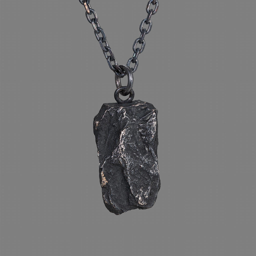 A dark, rugged silver pendant with a bold, chiseled texture and natural rock-like shape, hanging from a black oxidized chain against a plain gray background.