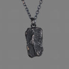 A dark, rugged silver pendant with a bold, chiseled texture and natural rock-like shape, hanging from a black oxidized chain against a plain gray background.