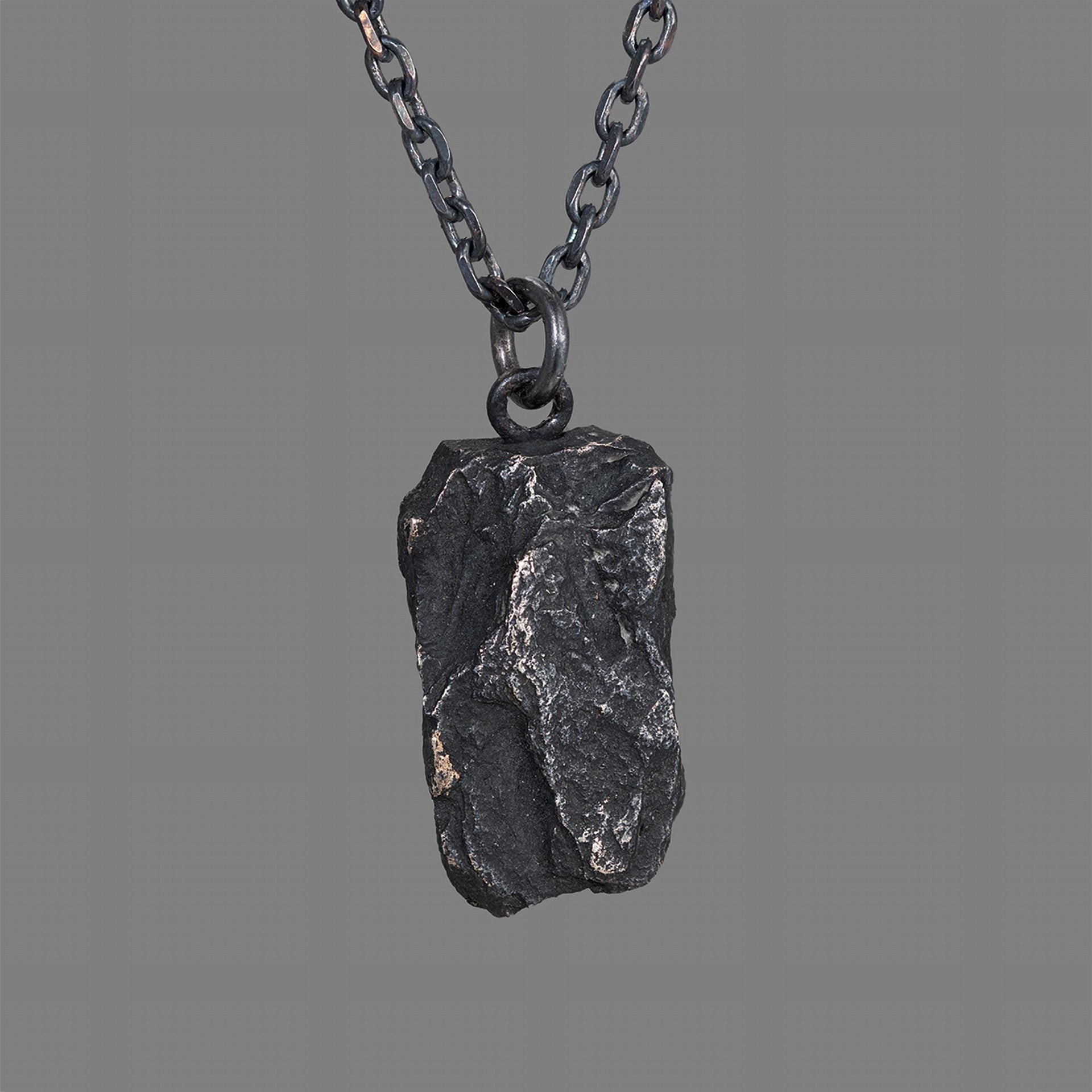 A dark, rugged silver pendant with a bold, chiseled texture and natural rock-like shape, hanging from a black oxidized chain against a plain gray background.