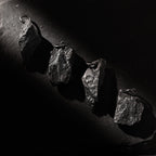 Four dark, rugged silver pendants shaped like natural rocks, displayed diagonally on a black textured surface. Each pendant has a unique shape and raw finish, highlighted by dramatic side lighting that casts deep shadows.