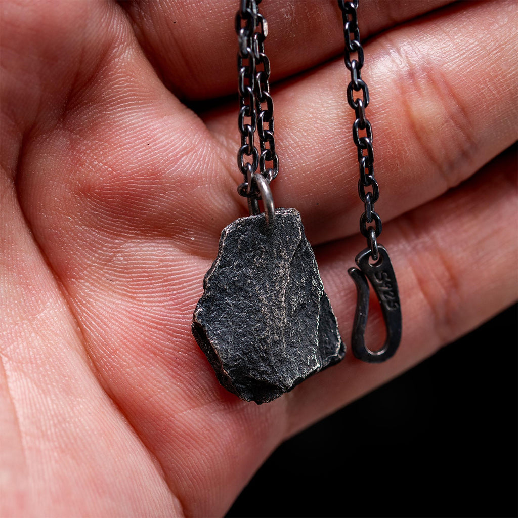 A dark, rugged silver pendant shaped like a natural rock, held by a hand. The pendant hangs from a black oxidized chain and features a raw, textured surface with an S925 stamped hook clasp.