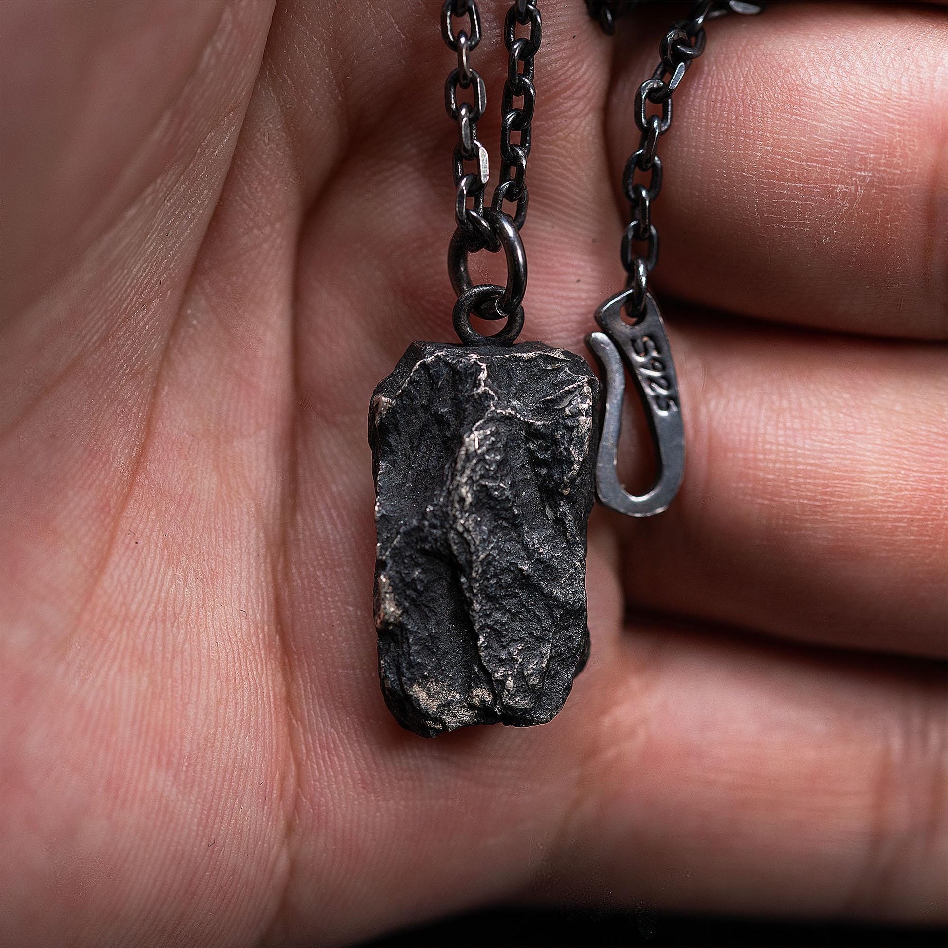 A dark, rugged silver pendant with a deeply rock-textured, layered surface, held between fingers. The pendant hangs from a black oxidized chain with an S925 stamped hook clasp, showcasing its natural rock-like form up close.