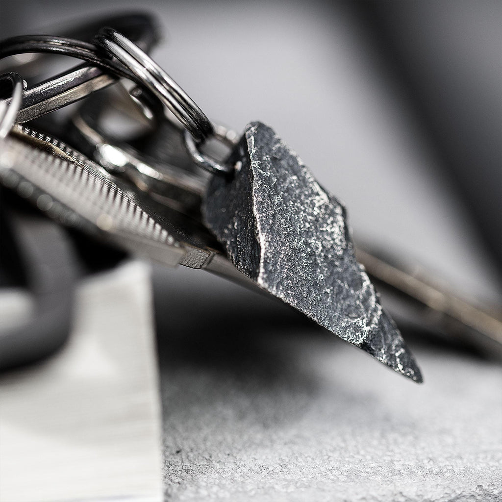 A dark, rugged silver pendant with a sharp, angular shape and gritty surface, attached to a keyring with multiple metal keys. The pendant rests on the edge of a closed book in a soft gray setting.