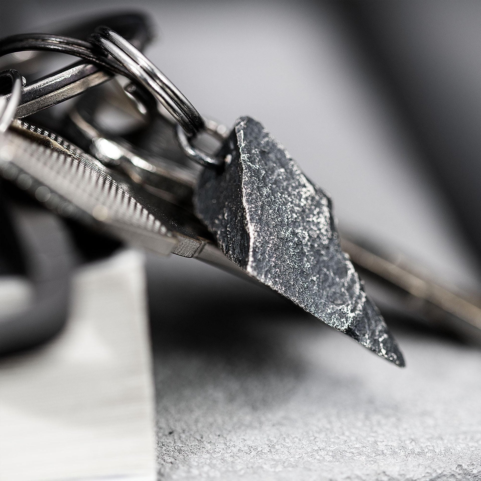 A dark, rugged silver pendant with a sharp, angular shape and gritty surface, attached to a keyring with multiple metal keys. The pendant rests on the edge of a closed book in a soft gray setting.