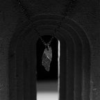 A dark, rugged silver pendant with a sharp, jagged shape, hanging from a black oxidized chain. The pendant is suspended in the center of a black stone archway, with dramatic shadows and lighting creating a moody, sculptural scene.