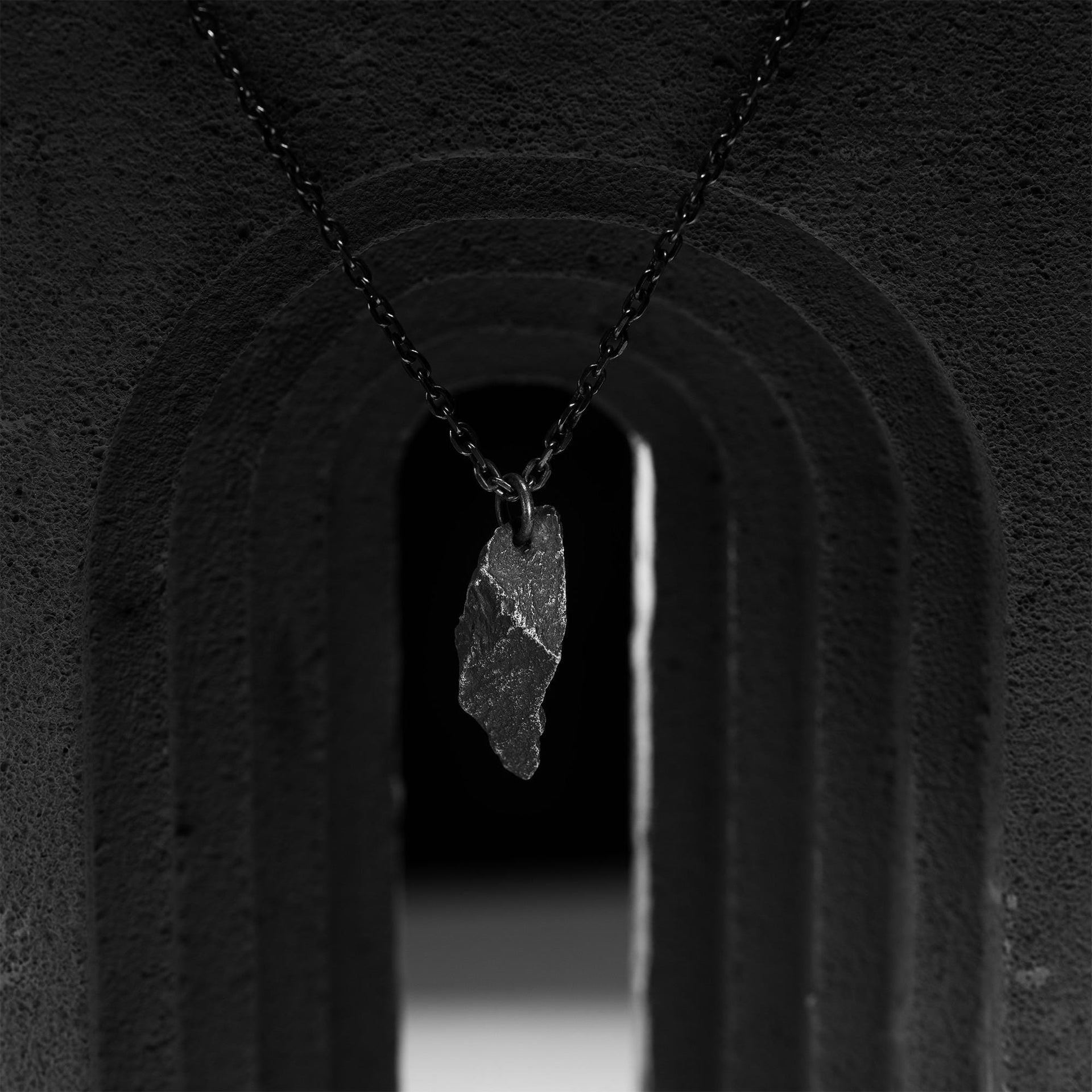 A dark, rugged silver pendant with a sharp, jagged shape, hanging from a black oxidized chain. The pendant is suspended in the center of a black stone archway, with dramatic shadows and lighting creating a moody, sculptural scene.