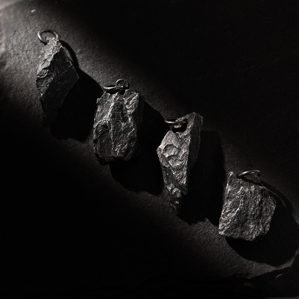 Four dark, rugged silver pendants shaped like natural rocks, displayed diagonally on a black textured surface. Each pendant has a unique shape and raw finish, highlighted by dramatic side lighting that casts deep shadows.