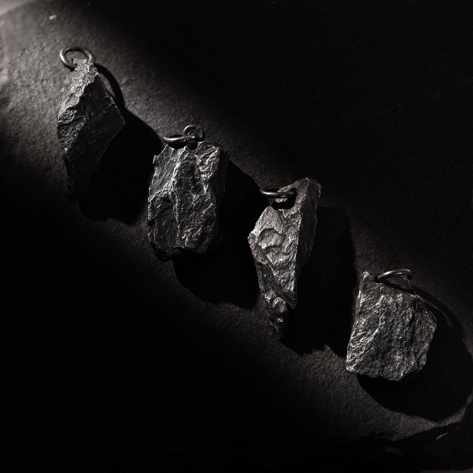 Four dark, rugged silver pendants shaped like natural rocks, displayed diagonally on a black textured surface. Each pendant has a unique shape and raw finish, highlighted by dramatic side lighting that casts deep shadows.