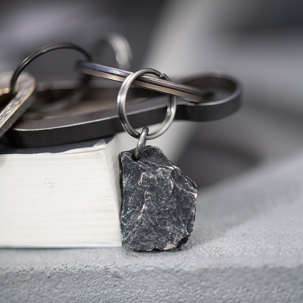 A dark, rugged silver pendant shaped like a natural rock, attached to a keyring with several metal keys and accessories. The pendant rests on the edge of a closed book with a soft grey background.
