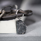 A dark, rugged silver pendant shaped like a natural rock, attached to a keyring with several metal keys and accessories. The pendant rests on the edge of a closed book with a soft grey background.