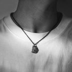 A dark, rugged silver pendant shaped like a natural rock, worn on a black oxidized chain around the neck. The pendant rests against a plain white T-shirt, shown in a black and white close-up of the chest area.