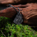 A dark, rugged silver pendant shaped like a natural rock, resting on green moss and leaning against a piece of textured driftwood. The pendant hangs from a black oxidized chain and features a raw, stone-like surface.