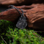 A dark, rugged silver pendant shaped like a natural rock, resting on green moss and leaning against a piece of textured driftwood. The pendant hangs from a black oxidized chain and features a raw, stone-like surface.