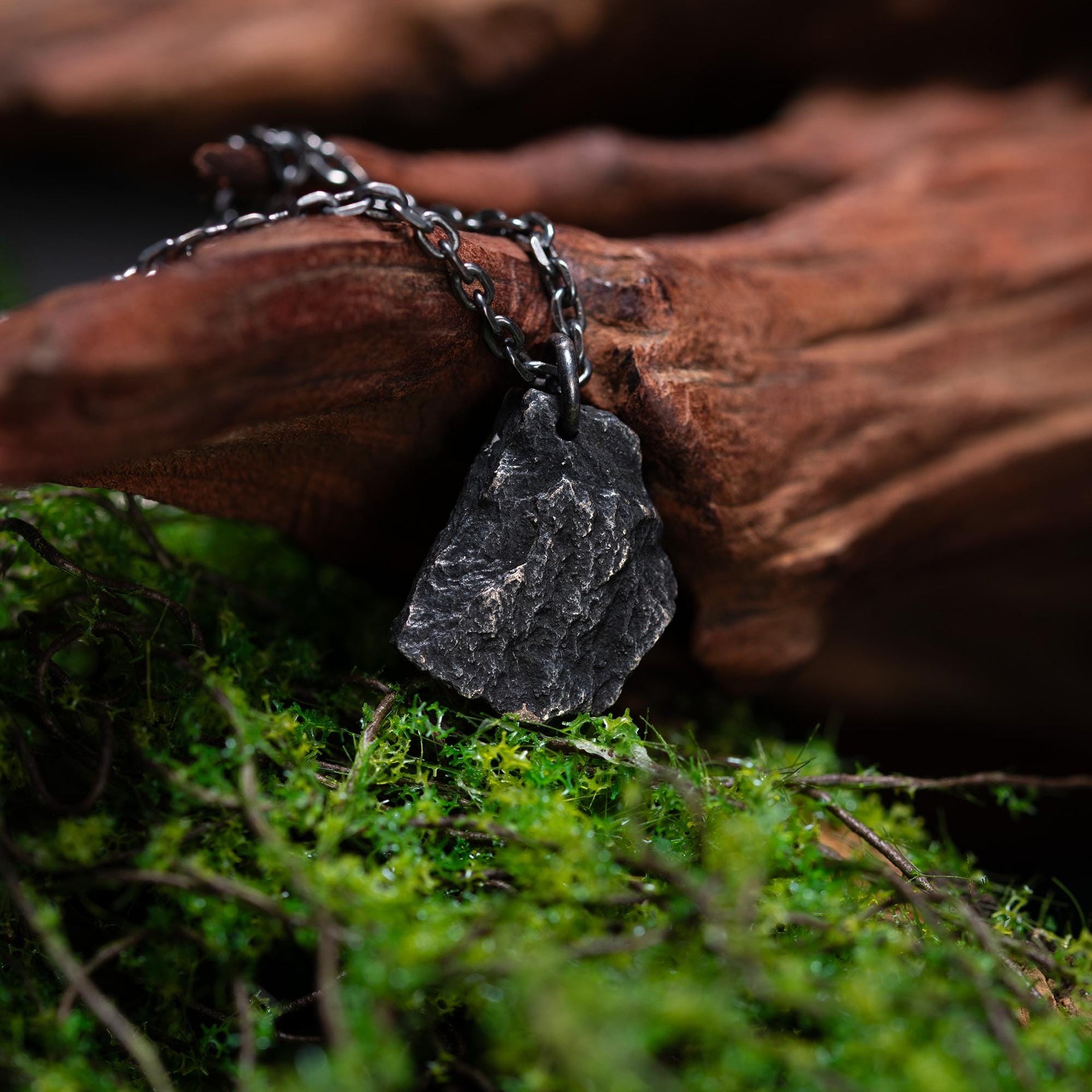 A dark, rugged silver pendant shaped like a natural rock, resting on green moss and leaning against a piece of textured driftwood. The pendant hangs from a black oxidized chain and features a raw, stone-like surface.