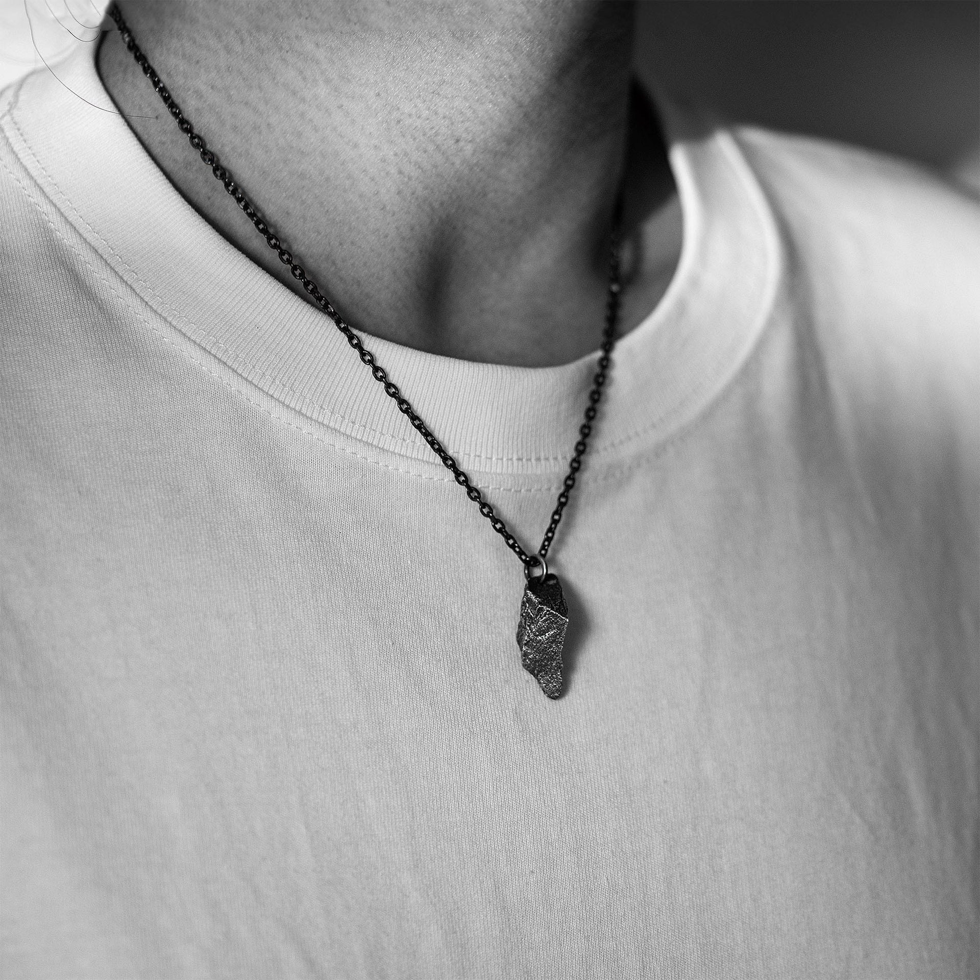 A dark, rugged silver pendant with a pointed, stone-like shape, worn on a black oxidized chain. The pendant rests on a plain white T-shirt in a black and white close-up of the upper chest and neckline.