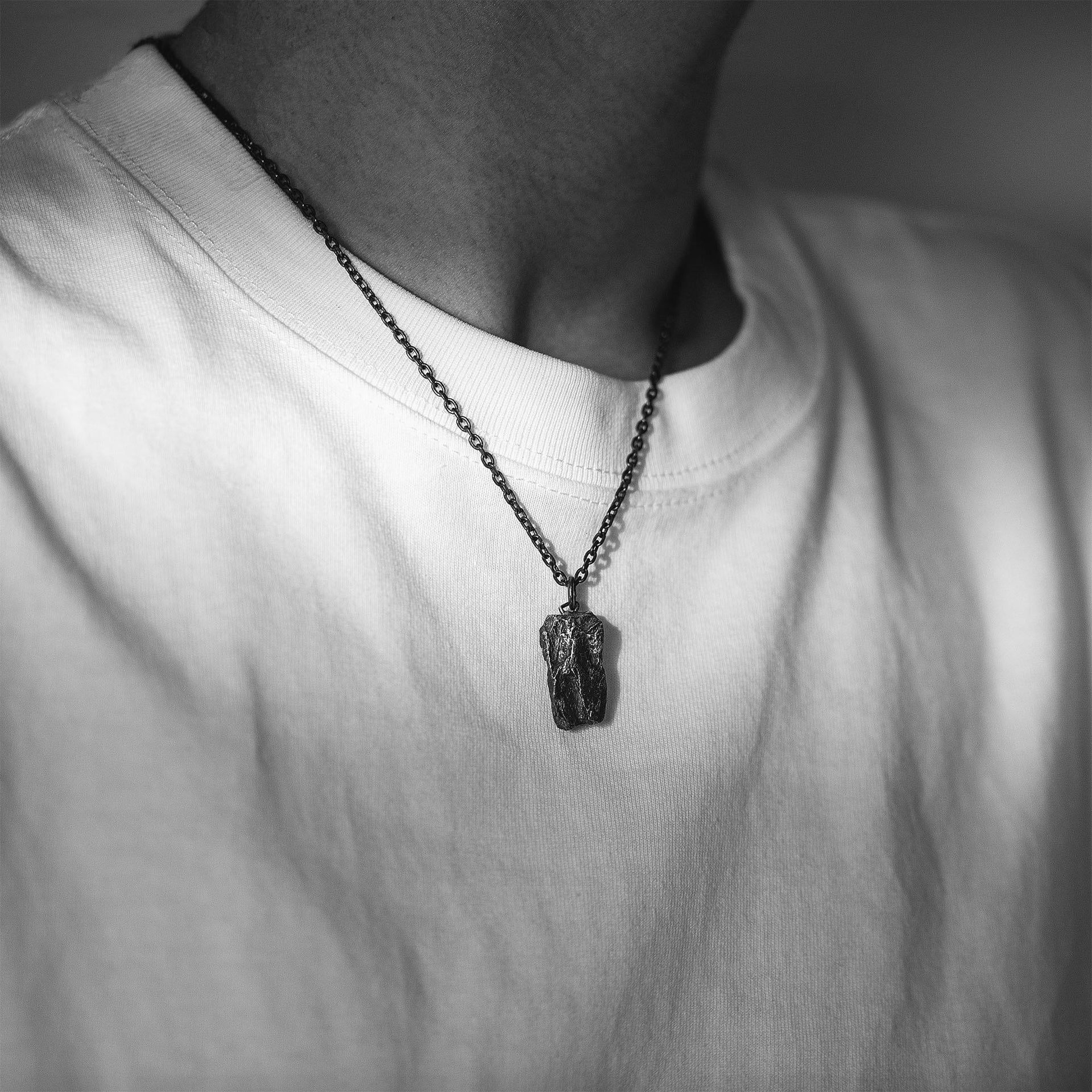 A dark, rugged silver pendant with a raw, layered surface, worn on a black oxidized chain around the neck. The pendant rests on a plain white T-shirt in a black and white close-up of the upper chest.