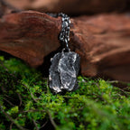 A dark, rugged silver pendant with a deeply layered and rock-textured surface, hanging from a black oxidized chain. The pendant rests on green moss and leans against a piece of weathered wood.