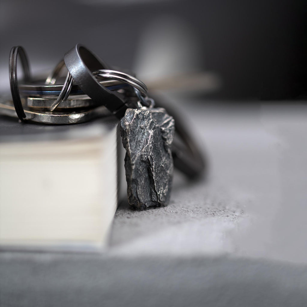 A dark, rugged silver pendant with a raw, layered surface, attached to a keyring resting on the edge of a closed book. The pendant hangs slightly over the book, with metal key loops in the background and a soft gray setting.