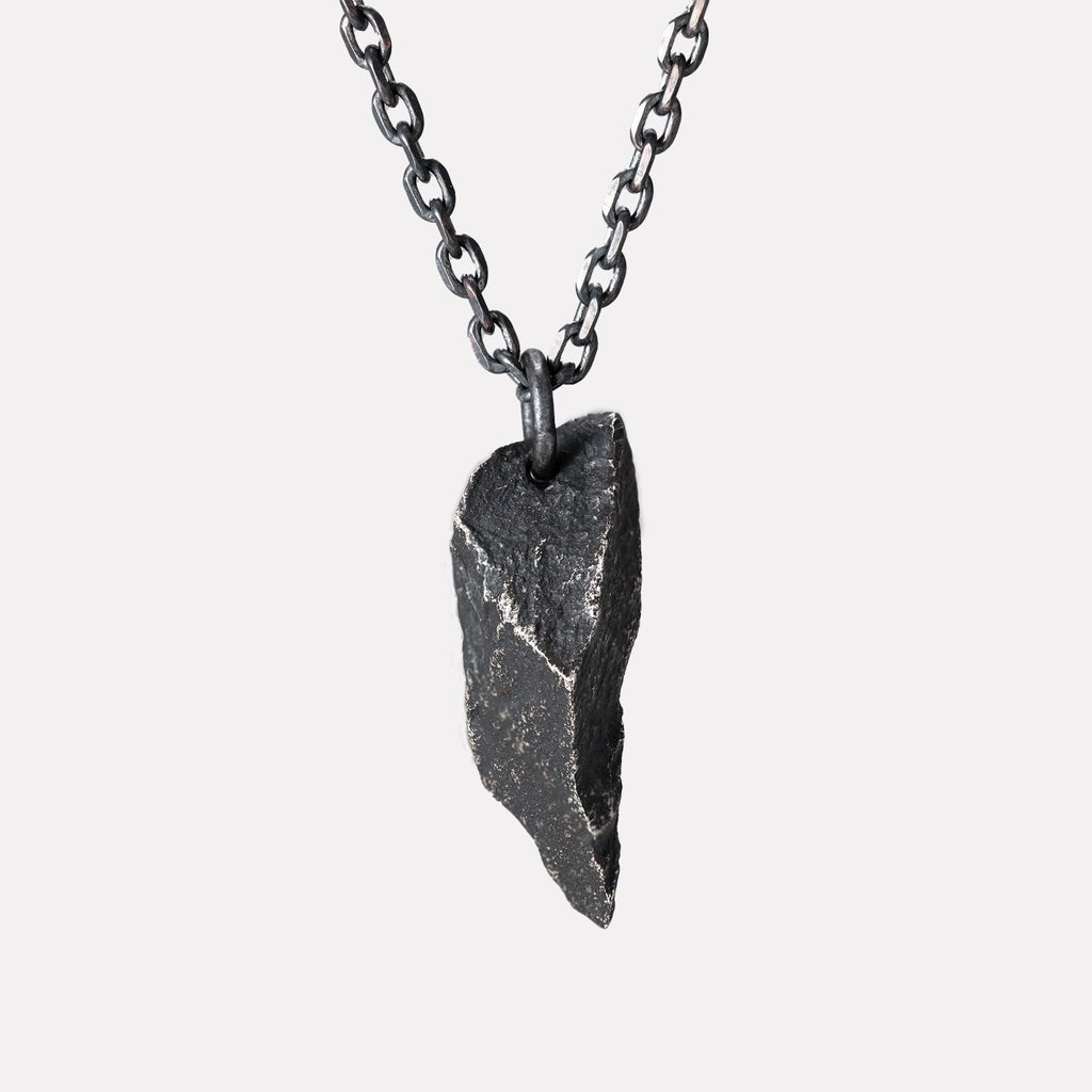 A dark, rugged silver pendant shaped like a sharp, angular rock with a rough, textured surface, hanging from a black oxidized chain against a plain white background.