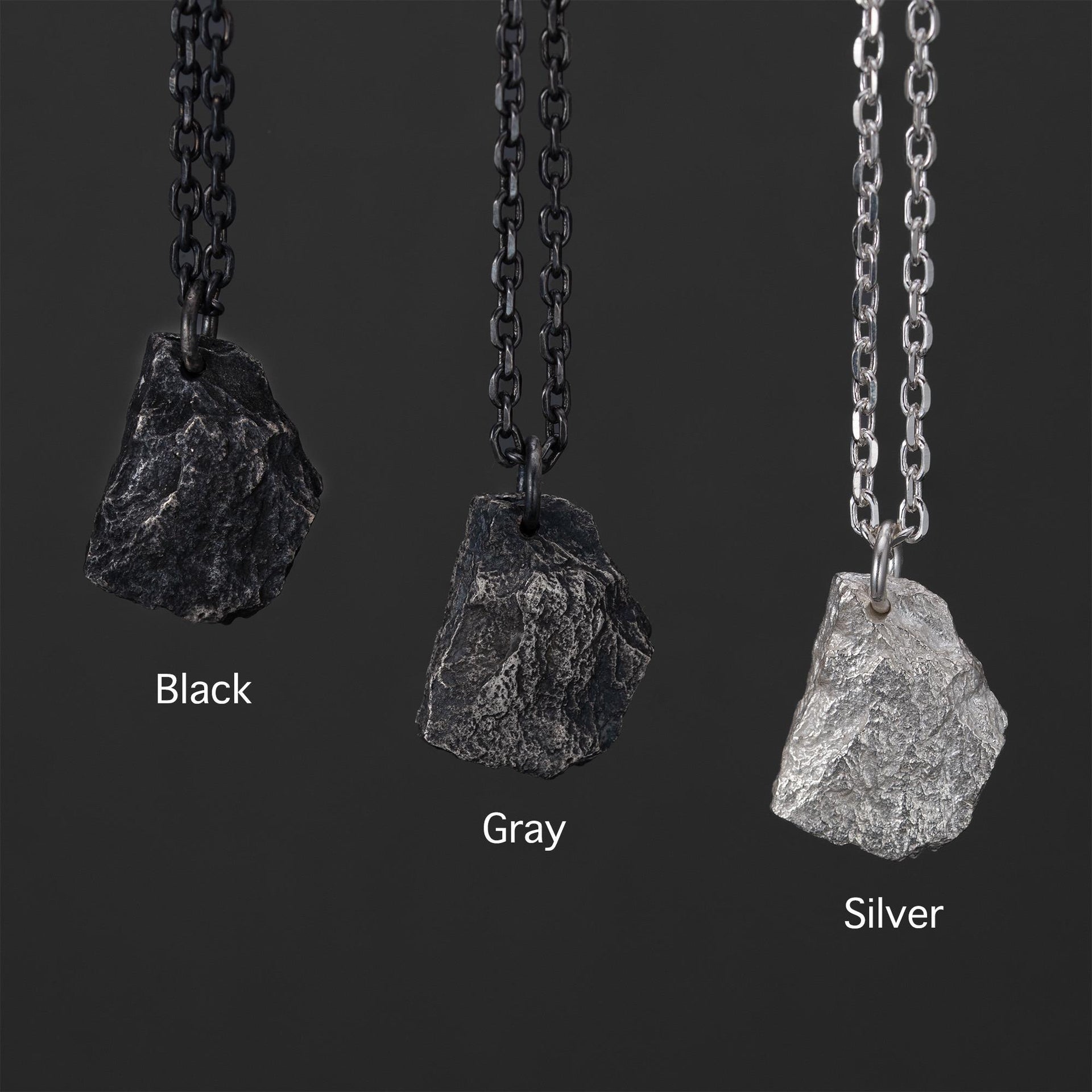 Three rugged silver pendants shaped like natural rocks, each hanging from a different chain. The finishes shown are Black with a dark chain, Gray with a muted chain, and Silver with a bright chain, displayed side by side on a dark background.