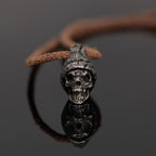 Beanie skull necklace standing on dark reflective glass with its mirror image, nickel silver hatted skull pendant on a leather cord